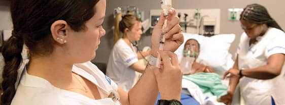 Three nursing students in uniform demonstrating assessment skills in a simulation lab.