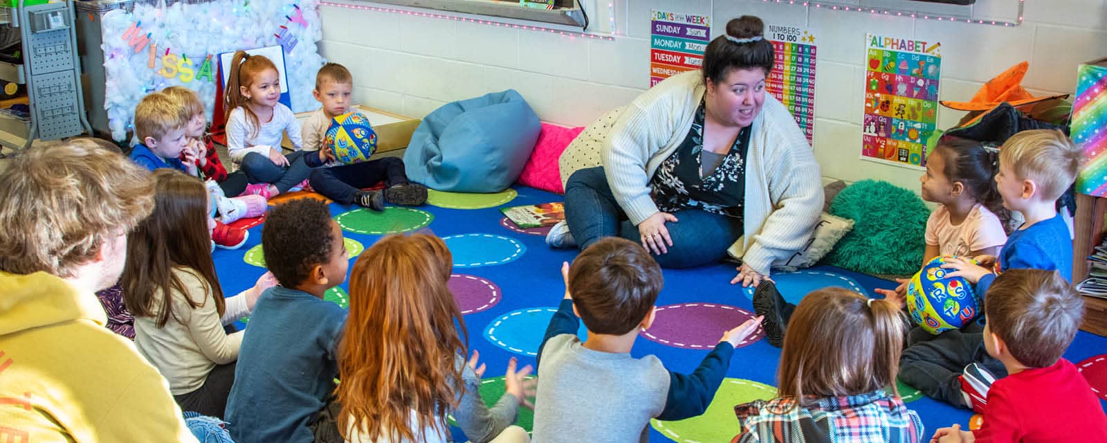 Several children sitting in a circle on a brightly colored carpet, engaging with the teacher during a large group activity.