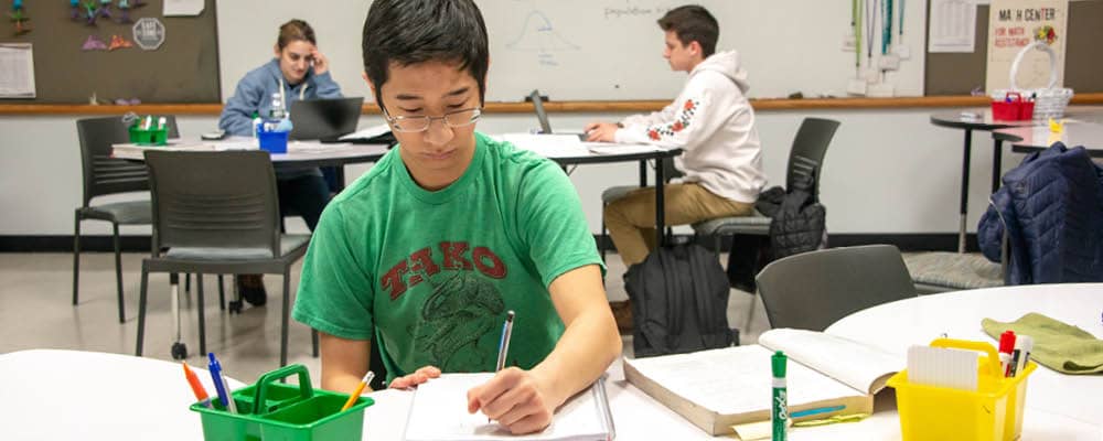 Three people studying in the FLCC Math Center. One student writes in a notebook while two others work on their laptops at nearby tables.